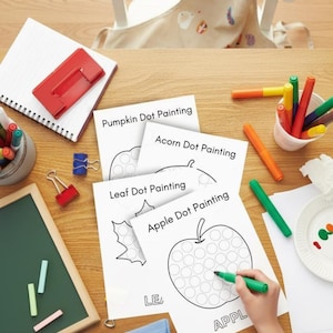 May include: A wooden table with art supplies and dot painting worksheets. The worksheets feature outlines of a pumpkin, acorn, leaf, and apple. A person is using a green marker to fill in the dots on the apple worksheet. Other supplies include markers, crayons, and a small chalkboard.