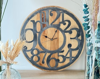 May include: A round wooden wall clock with a light brown face and dark gray numerals. The clock hands are black. The clock is surrounded by a dark gray border. The clock is displayed on a wooden surface.