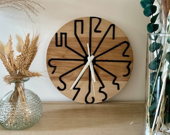 May include: A round wooden wall clock with black stylized numerals and hands. The clock face is light brown with a natural wood grain. The hands are white. The clock is displayed on a wooden shelf with decorative vases.