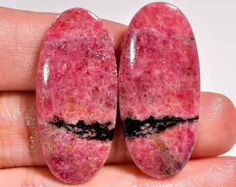 May include: Two polished, oval-shaped gemstones. The stones are a vibrant pink with a black band across the center. The gemstones are held in a hand, showcasing their smooth, reflective surfaces. The stones are likely used for jewelry making.