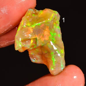 Könnte beinhalten: Ein roher Opal-Edelstein mit leuchtenden Farben von Grün, Orange und Gelb. Der Opal wird zwischen Fingern gehalten, was seine natürliche, unregelmäßige Form und sein schillerndes Farbspiel hervorhebt. Die Zahl "1" ist auf der oberen rechten Seite des Opals sichtbar.