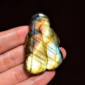 Può includere: Una pietra preziosa di labradorite lucidata con una forma organica unica. La pietra mostra un vibrante gioco di colori, con lampi di blu, oro e verde. La superficie è liscia e riflettente, mostrando i modelli e le inclusioni naturali all'interno della pietra.