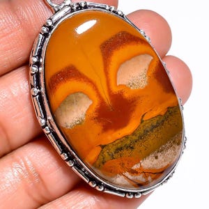 May include: Oval pendant featuring a polished, patterned gemstone in shades of orange, brown, and cream, set in a silver-toned metal frame. The stone's surface displays unique natural patterns.