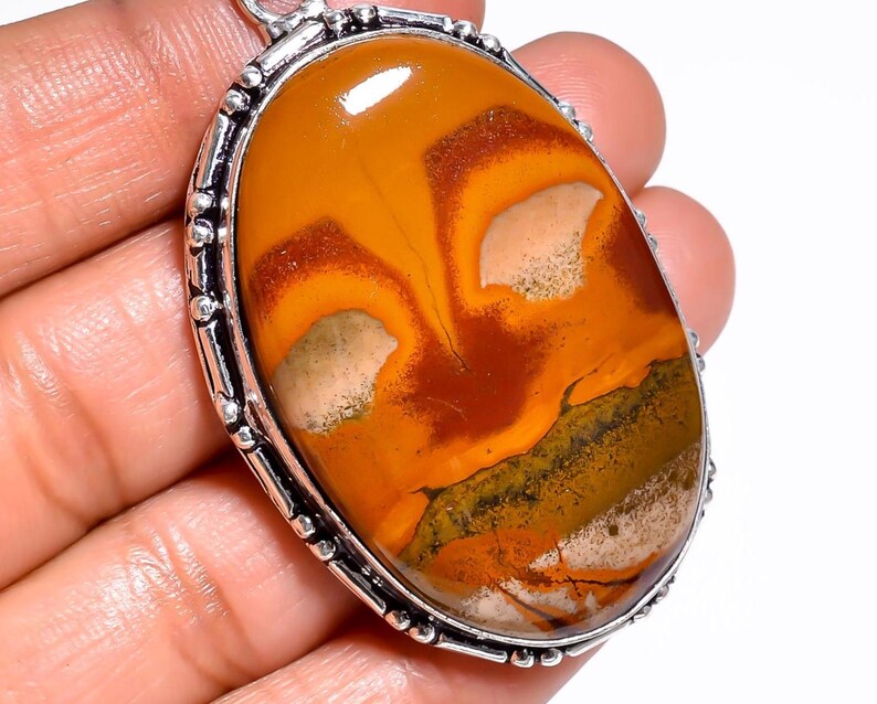 May include: Oval pendant featuring a polished, patterned gemstone in shades of orange, brown, and cream, set in a silver-toned metal frame. The stone's surface displays unique natural patterns.