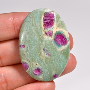 Puede incluir: Gema ovalada con una base de color verde claro, con inclusiones de rubí rosa vibrante. Los depósitos de rubí varían en tamaño y forma, creando un patrón único. La piedra tiene una superficie pulida, resaltando las texturas y colores naturales.