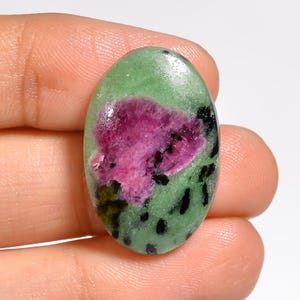 May include: An oval-shaped cabochon gemstone featuring a smooth, polished surface. The stone displays a vibrant mix of colours, primarily a light green base with a prominent, irregular magenta-pink inclusion in the centre and scattered black spots.