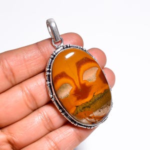 May include: An oval pendant with a silver-colored bail. The pendant features a polished, natural stone with a warm color palette of orange, brown, and cream. The stone is set in a silver-colored bezel with a beaded edge.