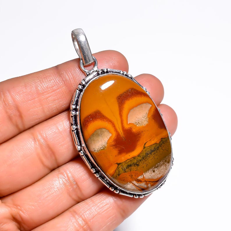 May include: An oval pendant with a silver-colored bail. The pendant features a polished, natural stone with a warm color palette of orange, brown, and cream. The stone is set in a silver-colored bezel with a beaded edge.