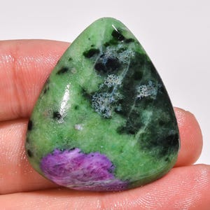 Peut inclure: Une pierre précieuse polie en forme de goutte d'eau avec une palette de couleurs vibrantes. La pierre présente une base verte, parsemée de motifs noirs et blancs, et une tache proéminente de violet profond. La gemme est tenue par une main.