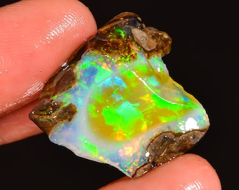 May include: A vibrant, raw opal gemstone with a mix of colors. The stone displays a play of color with bright green, blue, and yellow hues. The opal is held between fingers, showcasing its natural, rough texture and unique patterns.