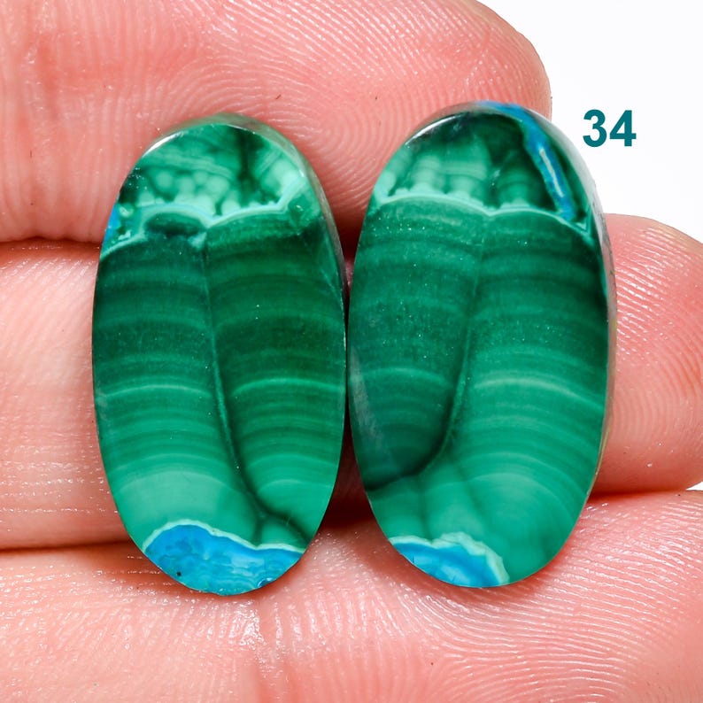 May include: Two oval-shaped green and blue gemstone cabochons with a banded pattern. The cabochons are set against a pink background. 34