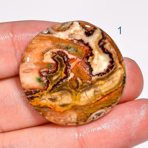May include: A round, polished gemstone with a swirling pattern of orange, brown, yellow, and white. The stone has a natural, earthy appearance, with visible variations in color and texture. The number "1" is visible in the upper right corner.