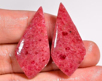 Puede incluir: Dos gemas pulidas en forma de lágrima, en tonos rosa y rojo. Las piedras tienen una superficie texturizada con pequeñas inclusiones rojas más oscuras. Las gemas se sostienen en una mano, mostrando su color y forma.