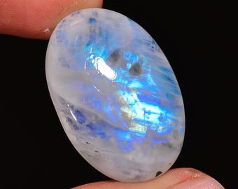 Può includere: Un cabochon ovale di pietra di luna bianca con un bagliore iridescente blu. La pietra ha una superficie liscia e lucida ed è lunga circa 5 cm.