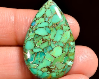 May include: A teardrop-shaped gemstone with a mosaic pattern of turquoise and gold-colored veins. The polished surface reflects light, highlighting the stone's natural beauty. The gemstone is held in a hand, showcasing its size and shape.