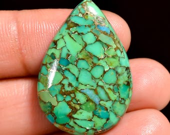 May include: A teardrop-shaped gemstone with a mosaic pattern of green, turquoise, and gold-colored veins. The polished surface of the stone reflects light, highlighting the intricate details of the natural design. The gemstone is held in a hand.