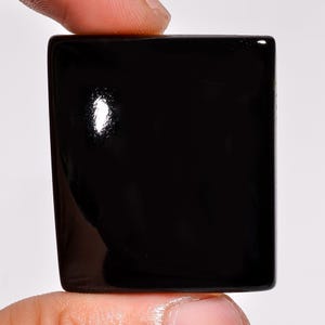 May include: A glossy, square-shaped black gemstone, held between two fingers. The stone has a polished surface with a slight curvature on the edges. The gemstone reflects light, creating highlights.