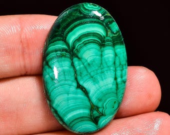 May include: Oval-shaped malachite gemstone with a polished surface. The stone features a vibrant green color with swirling patterns of lighter and darker green bands. The gemstone is held in a hand, showcasing its natural beauty.