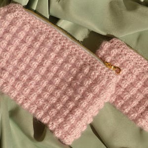 Op de afbeelding: Twee roze gehaakte etuis met gouden ritsen. De grotere etui is rechthoekig en de kleinere is cilindrisch. De haaksteek creëert een textuur met reliëf. De etuis worden gepresenteerd op een lichtgroene stof.