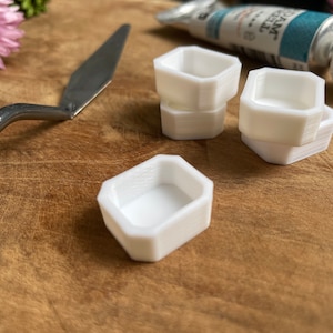 Könnte beinhalten: Vier weiße Plastik-Farbpaletten, jede mit einer sechseckigen Form und einer flachen Vertiefung, sind auf einer Holzoberfläche angeordnet. Ein silbernes Metall-Palette-Messer ist ebenfalls auf dem Bild zu sehen.