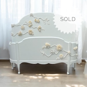 May include: A white bed frame with floral and bird embellishments. The headboard and footboard feature carved details and decorative cream-colored flowers and vines. The word "SOLD" is displayed on the headboard.