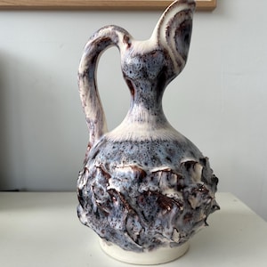 Peut inclure: Pichet en céramique au design texturé unique. Le pichet présente un corps bulbeux avec une surface texturée et épineuse, et une fine poignée. La palette de couleurs comprend des nuances de blanc, de bleu et de marron, créant une pièce visuellement intéressante.