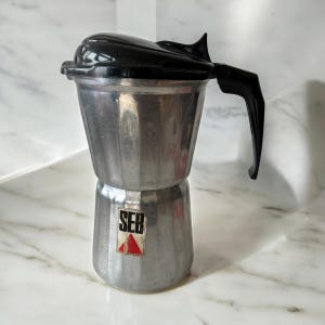 May include: A vintage silver-toned stovetop espresso maker with a black handle and lid. The coffee pot has a classic design with a SEB logo on the front. A practical kitchen item for brewing coffee.