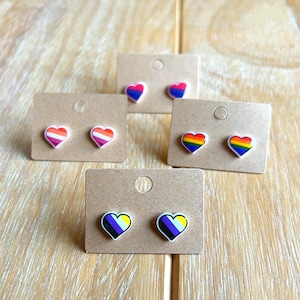 May include: Four pairs of heart-shaped earrings with different pride flag designs. The earrings are displayed on small brown cards. The flags are: lesbian, bisexual, transgender, and non-binary.
