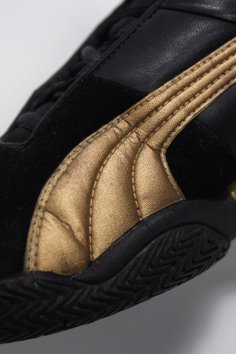K&ouml;nnte beinhalten: Schwarze und goldene Sneaker mit einem Obermaterial aus Wildleder und einer strukturierten Gummisohle.