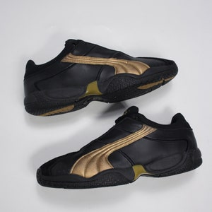 K&ouml;nnte beinhalten: Ein Paar schwarze Lederschuhe mit goldenen Akzenten. Die Schuhe haben einen niedrigen Schaft und eine Gummisohle.