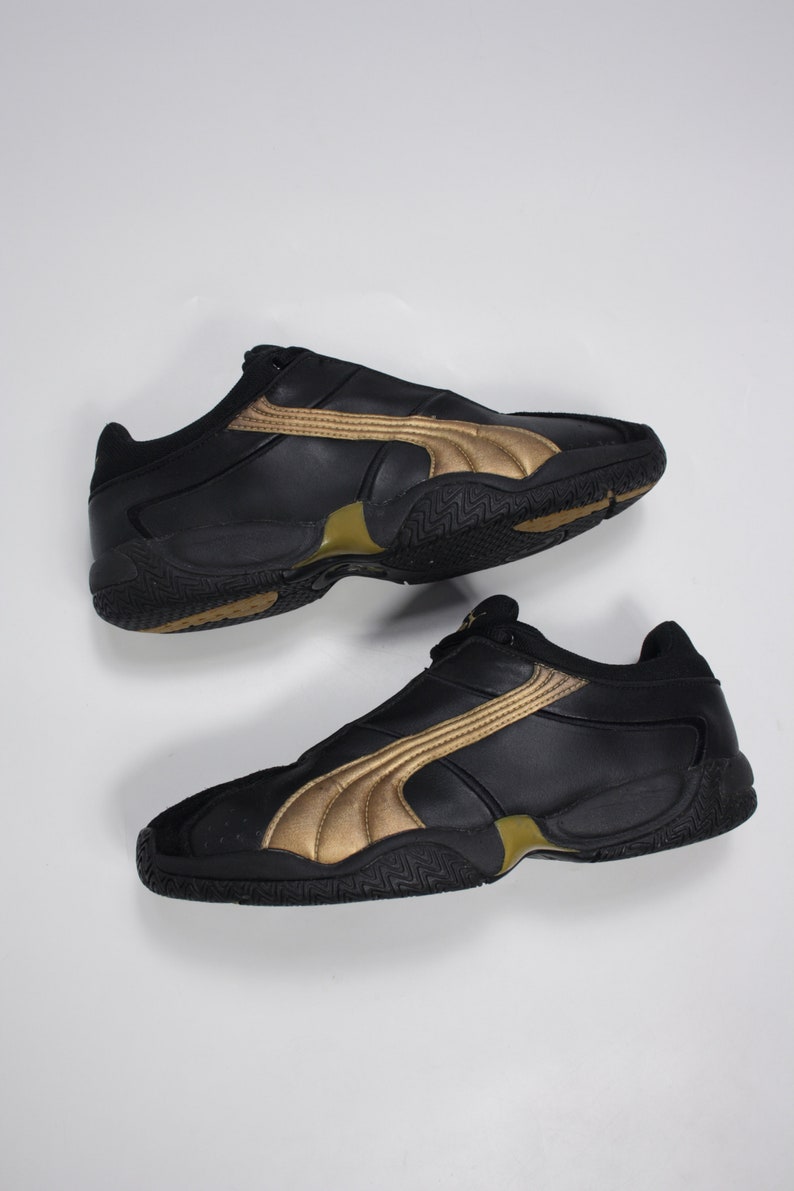 K&ouml;nnte beinhalten: Ein Paar schwarze Lederschuhe mit goldenen Akzenten. Die Schuhe haben einen niedrigen Schaft und eine Gummisohle.
