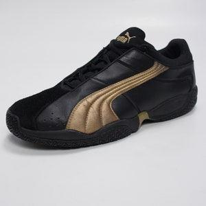 K&ouml;nnte beinhalten: Schwarze Lederschuhe mit goldenem Puma-Logo und goldenen Akzenten. Die Schuhe haben eine schwarze Gummisohle mit einem strukturierten Muster.