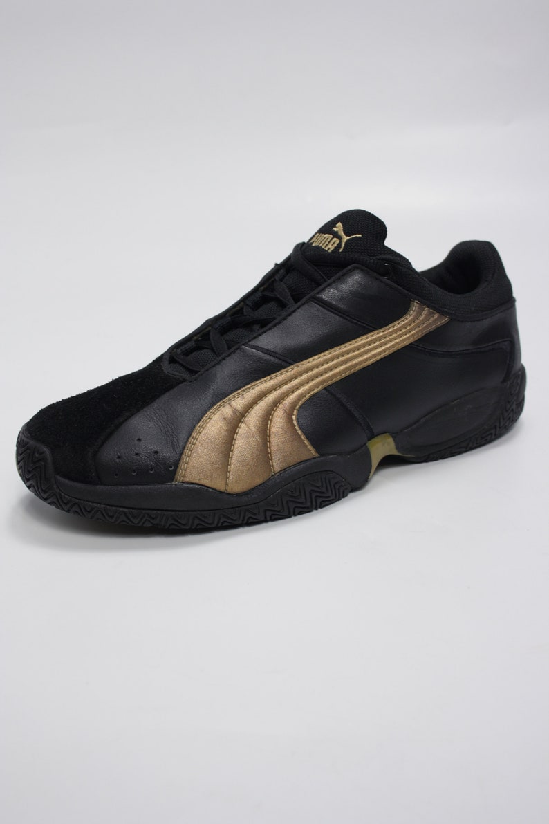 K&ouml;nnte beinhalten: Schwarze Lederschuhe mit goldenem Puma-Logo und goldenen Akzenten. Die Schuhe haben eine schwarze Gummisohle mit einem strukturierten Muster.