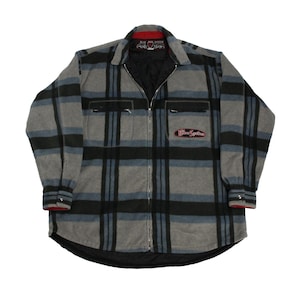 Puede incluir: Una camisa de franela a cuadros gris, azul y negro con cierre de cremallera. La camisa tiene un forro negro y un parche rojo y negro en el cuello que dice "Blue Jean Spirit".