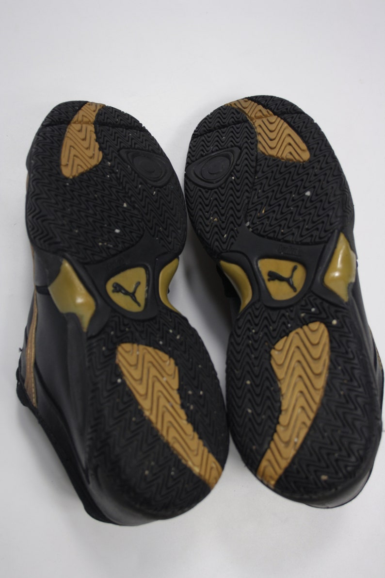 K&ouml;nnte beinhalten: Ein Paar schwarze und goldene Sportschuhe mit einer strukturierten Sohle. Die Schuhe haben ein schwarzes Leder-Obermaterial und einen goldenen Rand um die Sohle.