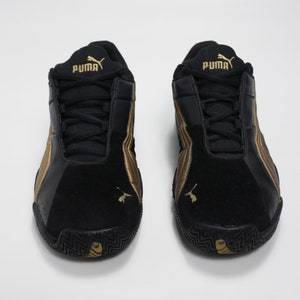 K&ouml;nnte beinhalten: Ein Paar schwarze und goldene Puma-Sneaker mit Wildledereinsatz und Gummisohle.