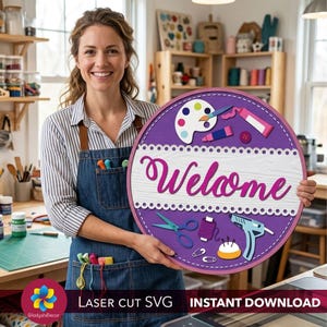 Puede incluir: Un letrero redondo de madera, morado y blanco, con la palabra "Welcome" en escritura rosa. El letrero presenta ilustraciones temáticas de artesanía, incluyendo una paleta de pintura, tijeras y una pistola de pegamento. Una persona sostiene el letrero, usando un delantal de mezclilla.
