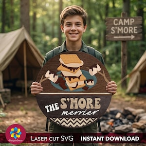 May include: A wooden sign with a s'mores design, featuring graham crackers, chocolate, and marshmallows. The sign reads "The S'MORE merrier" and is ideal for a camping theme. The sign is held by a person in a forest setting.