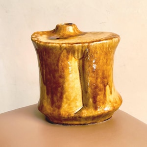 Può includere: Un vaso in ceramica con una smaltatura lucida e maculata nei toni del giallo senape e del marrone. Il recipiente presenta una forma cilindrica organica e irregolare con un'apertura stretta decentrata sulla parte superiore e una base svasata.