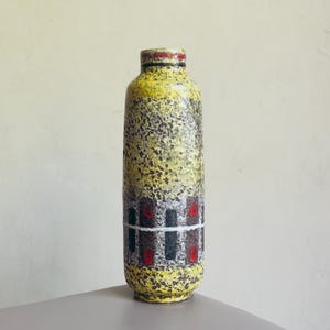 May include: A tall, cylindrical ceramic vase with a textured surface. The vase has a yellow and brown speckled pattern, with a band of black, red, and white geometric shapes. The rim is red and the base is yellow.