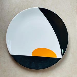 May include: A round, white ceramic plate with a modern geometric design. The plate features a black and white colour scheme with an orange semi-circle and a small green triangle. The plate is suitable for serving food.