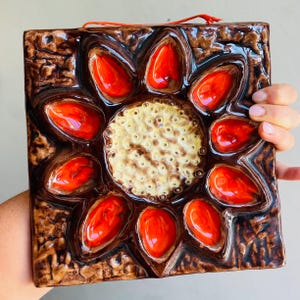 May include: Square ceramic wall art with a floral design. The artwork features a central cream-colored circle surrounded by ten vibrant red petal-shaped elements. The background is a textured brown, and a red string is attached for hanging.