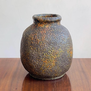 Puede incluir: Un jarrón de cerámica redondo con una superficie gruesa y texturizada que presenta un color base gris carbón oscuro con parches moteados de naranja quemado y amarillo mostaza. El recipiente tiene un cuerpo ancho y bulboso y un borde corto y grueso.