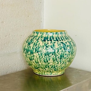 May include: A round ceramic vase with a speckled green and cream pattern. The interior rim is yellow. The vase sits on a gray surface against a white wall.