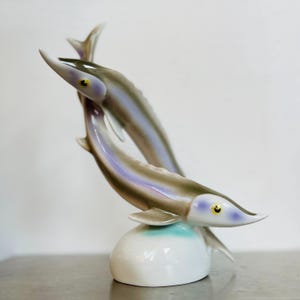 Könnte beinhalten: Eine Porzellanfigur aus zwei stilisierten Stören, wobei einer über den anderen springt. Die Fische sind in Grautönen, Lila und Weiß bemalt, mit gelben Augen. Der Sockel ist weiß mit einem hellblauen Akzent.