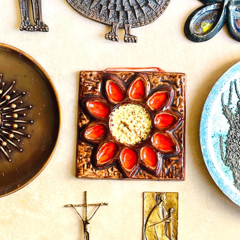 May include: A collection of vintage decorative items, including a square ceramic tile with a sunflower design in red and yellow, a bronze plate, and other metal art pieces. The items are arranged on a light-colored surface.