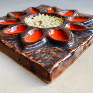 May include: A square, ceramic wall tile with a raised floral design. The tile features a dark brown base with a flower-like pattern of orange and cream-colored accents. The tile has a glossy finish and a textured surface.