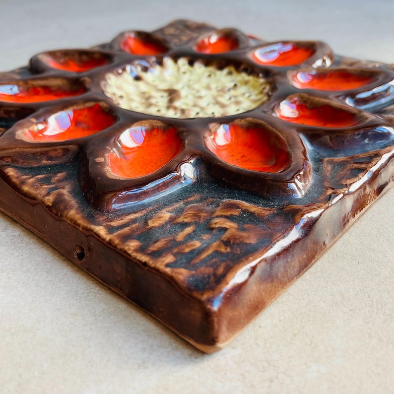 May include: A square, ceramic wall tile with a raised floral design. The tile features a dark brown base with a flower-like pattern of orange and cream-colored accents. The tile has a glossy finish and a textured surface.