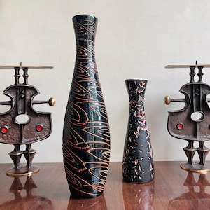 May include: Two tall ceramic vases and two bronze-toned metal candle holders. The taller vase is black with orange swirling patterns. The shorter vase is black with textured red markings. The two metal candle holders feature abstract shapes.