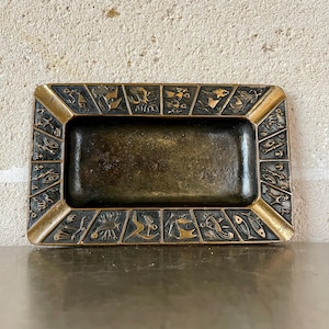 Könnte beinhalten: Rechteckiger Aschenbecher in Bronzefarbe mit dekorativem Rand. Der Rand weist eine Reihe von geprägten, stilisierten Figuren und Symbolen in einem sich wiederholenden Muster auf. Der Aschenbecher hat eine vertiefte Mitte und einen goldfarbenen Rand.